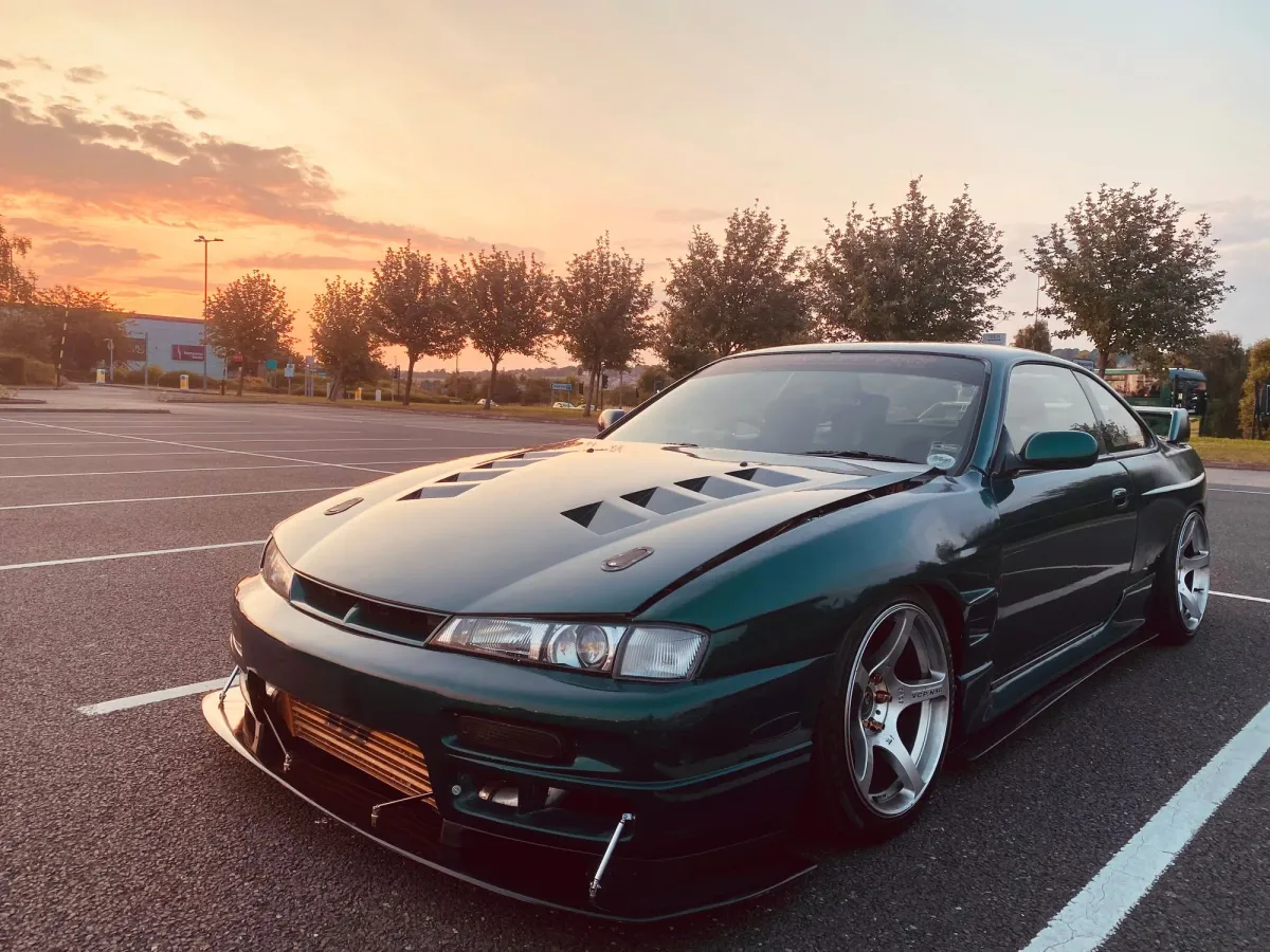 Dark green Nissan S14 Kouki with the sharper facelift headlights, a vented bonnet and a front splitter with support rods, in a car park at sunset